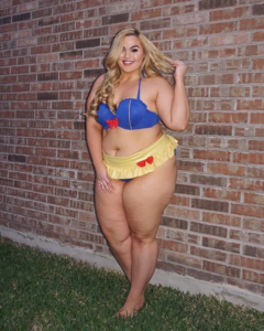 Full figured blonde woman in bra and panties, standing against a brick wall smiling.
