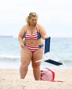 Chubby blonde in patriotic bikini on the beach carrying a beach chair.